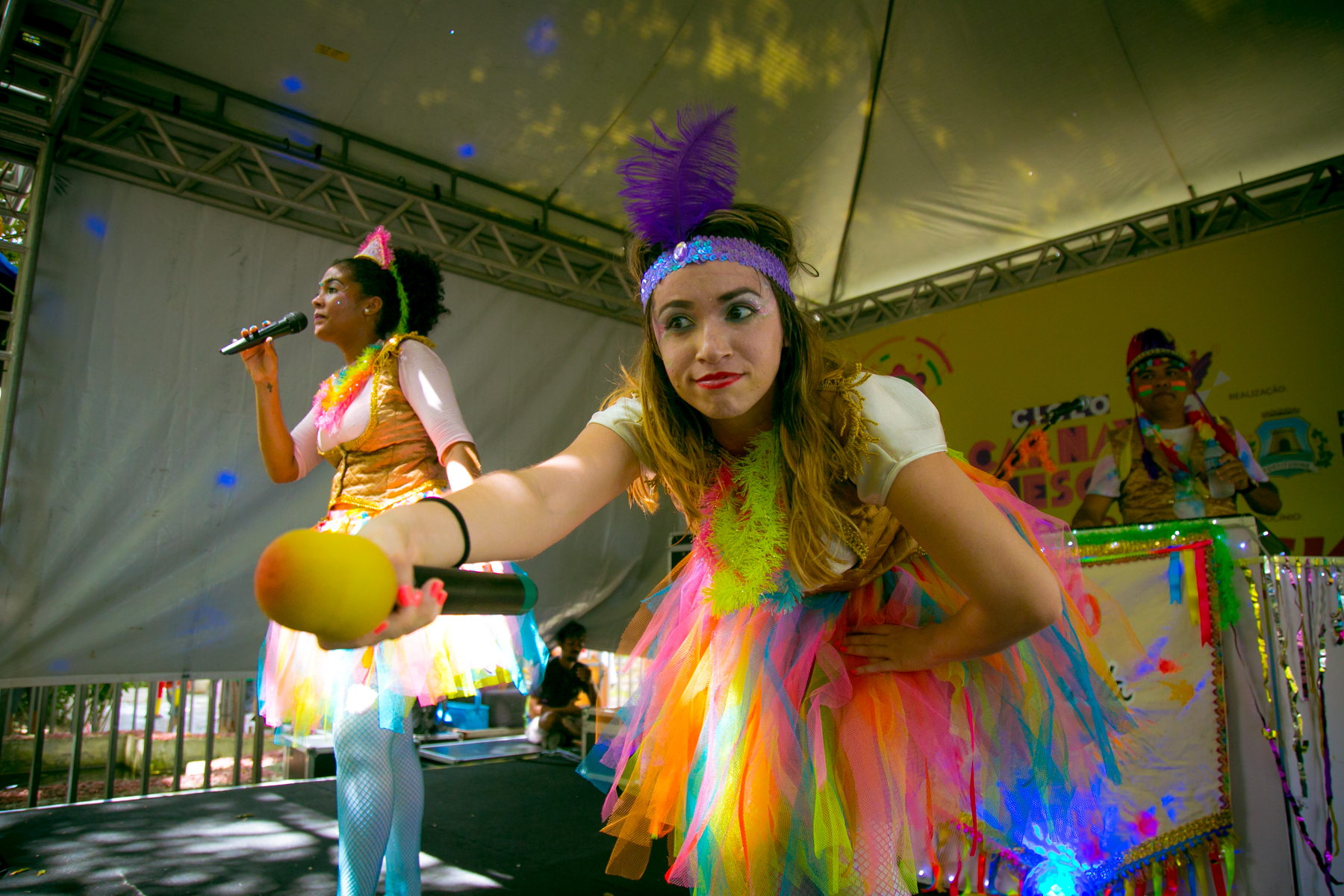 Dua integrantes do Trio Aquarela  dançando vestindo roupas coloridas. Uma delas direciona o microfone para o público.